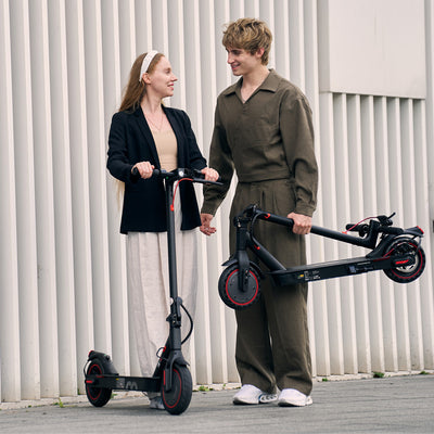 Two people standing with electric scooters against a light-colored wall.